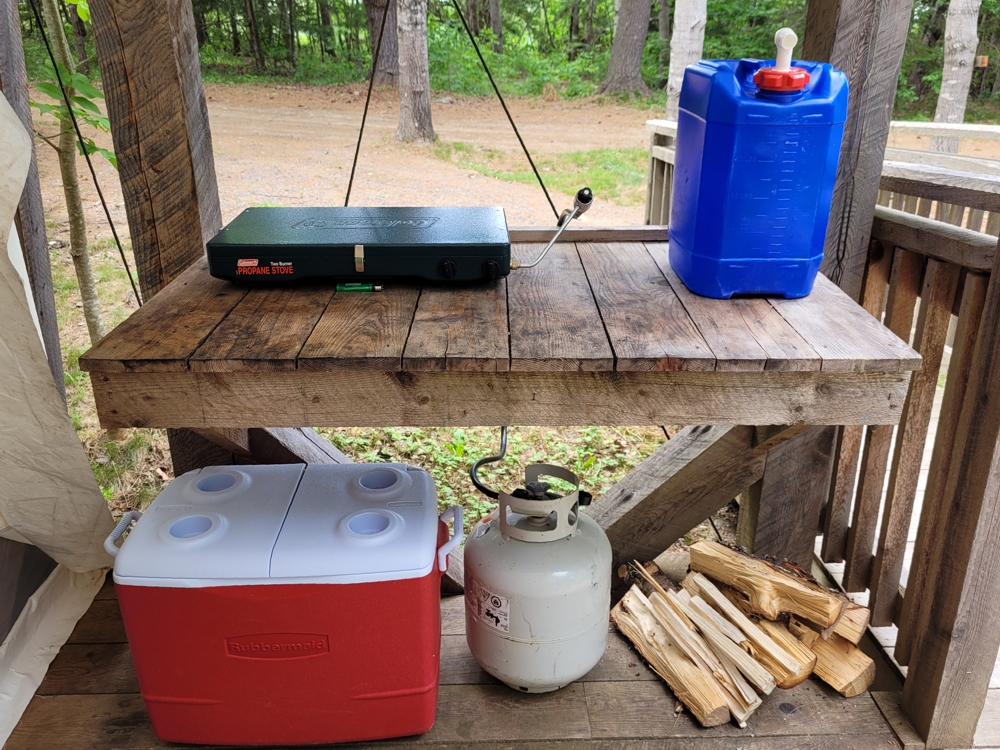 Glamping tent outdoor cooking setup with propane stove, cooler, water container and firewood in Nova Scotia